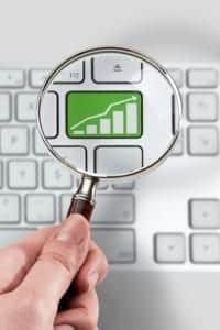 A hand holding a magnifying glass zooms in on a green button on a keyboard with a chart showing financial growth